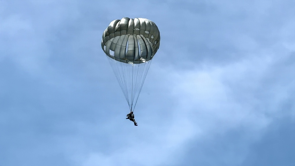 A single parachutist is descending through a partly cloudy blue sky using a round, olive-drab/gray military-style parachute. The canopy is fully deployed and appears to be in stable descent. The image is taken from a ground-level upward perspective, silhouetting the jumper against the sky.