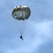 A single parachutist is descending through a partly cloudy blue sky using a round, olive-drab/gray military-style parachute. The canopy is fully deployed and appears to be in stable descent. The image is taken from a ground-level upward perspective, silhouetting the jumper against the sky.