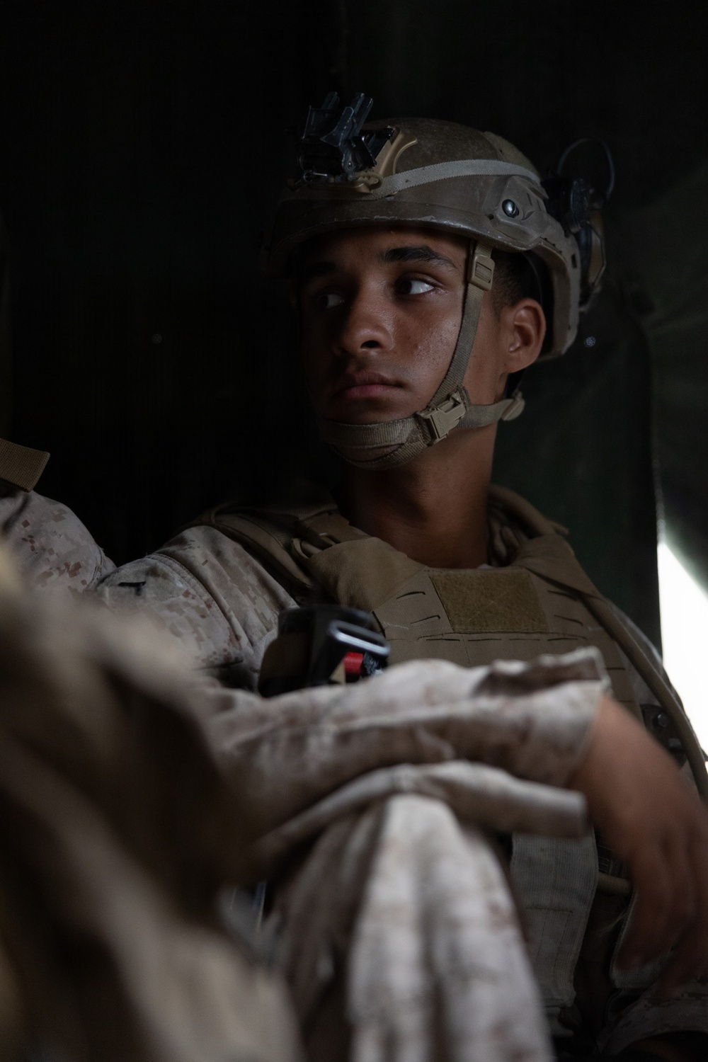 A young male military service member is seated, wearing full combat gear including a tactical helmet with night vision device mount, body armor, and desert camouflage (MARPAT) uniform. He is gazing upward and to the side with a focused, contemplative expression. The image appears to be taken inside a vehicle or aircraft, with dramatic low-key lighting emphasizing his face.