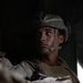 A young male military service member is seated, wearing full combat gear including a tactical helmet with night vision device mount, body armor, and desert camouflage (MARPAT) uniform. He is gazing upward and to the side with a focused, contemplative expression. The image appears to be taken inside a vehicle or aircraft, with dramatic low-key lighting emphasizing his face.