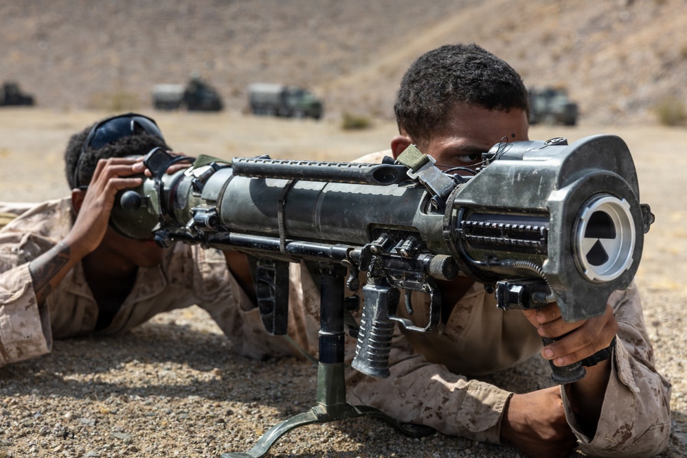 Two military personnel in desert camouflage uniforms are in a prone firing position in an arid, desert training environment. The foreground soldier is aiming a recoilless rifle or anti-tank weapon mounted on a bipod. Military vehicles are visible in the blurred background, suggesting a live-fire training exercise.