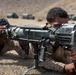 Two military personnel in desert camouflage uniforms are in a prone firing position in an arid, desert training environment. The foreground soldier is aiming a recoilless rifle or anti-tank weapon mounted on a bipod. Military vehicles are visible in the blurred background, suggesting a live-fire training exercise.