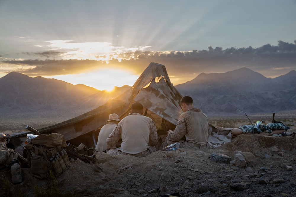 Three military personnel in desert camouflage uniforms are seated on rocky elevated ground facing a dramatic sunset over a mountain range. They appear to be at a field position with a makeshift camouflage shelter or tarp structure behind them. Military gear, tactical vests, and equipment are visible nearby.