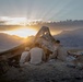 Three military personnel in desert camouflage uniforms are seated on rocky elevated ground facing a dramatic sunset over a mountain range. They appear to be at a field position with a makeshift camouflage shelter or tarp structure behind them. Military gear, tactical vests, and equipment are visible nearby.