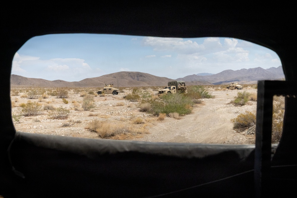 The image is taken from inside a military vehicle looking out through a rear window at a desert landscape. Three military vehicles, appearing to be HMMWVs (Humvees), are positioned on a dirt trail in an arid, scrub-covered desert terrain with mountain ranges visible in the background. The scene suggests a military convoy or patrol operation in a desert training or operational environment.