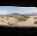 The image is taken from inside a military vehicle looking out through a rear window at a desert landscape. Three military vehicles, appearing to be HMMWVs (Humvees), are positioned on a dirt trail in an arid, scrub-covered desert terrain with mountain ranges visible in the background. The scene suggests a military convoy or patrol operation in a desert training or operational environment.