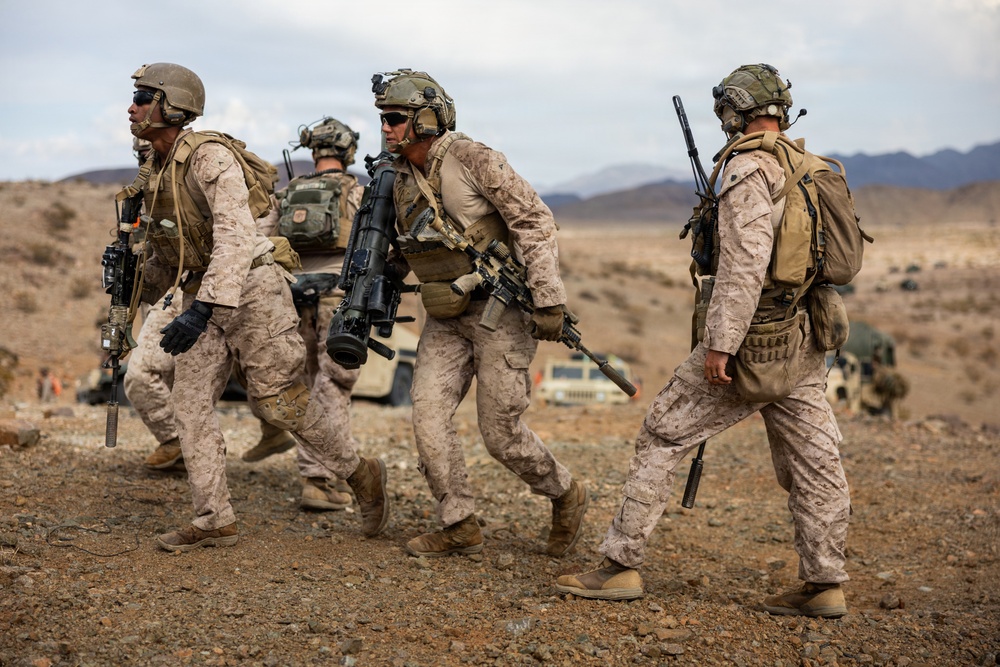 A group of four military personnel in desert camouflage uniforms are moving across arid terrain in what appears to be a training exercise or patrol operation. They are fully equipped with tactical gear, helmets, body armor, and various weapons including rifles and what appears to be a rocket launcher or recoilless rifle. Military vehicles, including Humvees, are visible in the background against a desert mountain landscape.