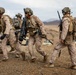 A group of four military personnel in desert camouflage uniforms are moving across arid terrain in what appears to be a training exercise or patrol operation. They are fully equipped with tactical gear, helmets, body armor, and various weapons including rifles and what appears to be a rocket launcher or recoilless rifle. Military vehicles, including Humvees, are visible in the background against a desert mountain landscape.