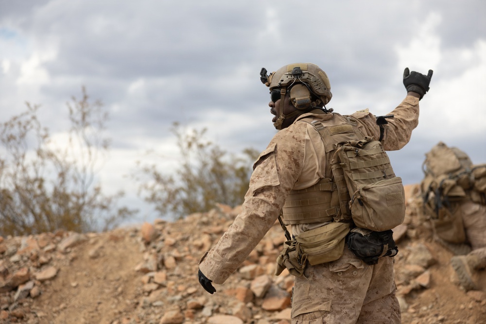 A soldier in desert camouflage (MARPAT pattern) is photographed from behind, raising one hand in what appears to be a hand signal gesture. The terrain is rocky and arid, consistent with a desert training or operational environment. A second partially visible soldier can be seen in the background to the right.