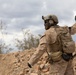 A soldier in desert camouflage (MARPAT pattern) is photographed from behind, raising one hand in what appears to be a hand signal gesture. The terrain is rocky and arid, consistent with a desert training or operational environment. A second partially visible soldier can be seen in the background to the right.