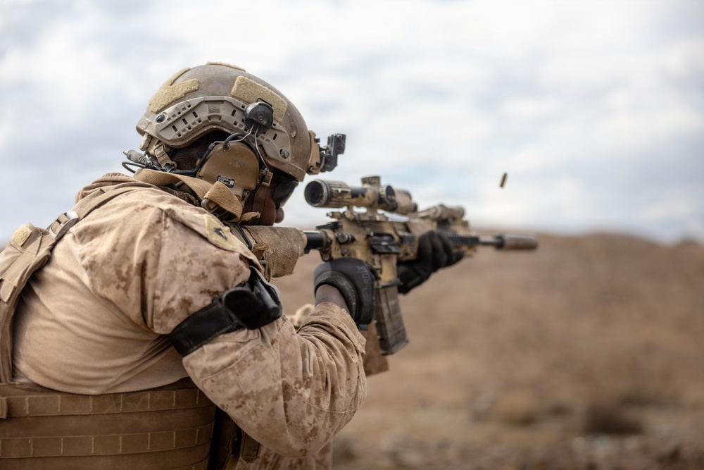 A soldier in desert camouflage uniform is actively firing a rifle, with a spent brass casing visibly ejecting from the weapon. The individual is equipped with full tactical gear including a combat helmet with mounted accessories, body armor, and tactical gloves. The setting appears to be an arid, desert-like environment under an overcast sky.
