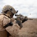 A soldier in desert camouflage uniform is actively firing a rifle, with a spent brass casing visibly ejecting from the weapon. The individual is equipped with full tactical gear including a combat helmet with mounted accessories, body armor, and tactical gloves. The setting appears to be an arid, desert-like environment under an overcast sky.
