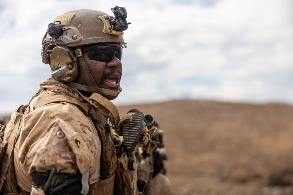 A military service member in full combat gear is photographed in a desert or arid environment, appearing to be shouting or issuing commands. The soldier is equipped with modern tactical equipment including a helmet-mounted device, communication headset, body armor, and tactical vest. The background shows a barren, hilly landscape under a partly cloudy sky.