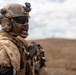 A military service member in full combat gear is photographed in a desert or arid environment, appearing to be shouting or issuing commands. The soldier is equipped with modern tactical equipment including a helmet-mounted device, communication headset, body armor, and tactical vest. The background shows a barren, hilly landscape under a partly cloudy sky.