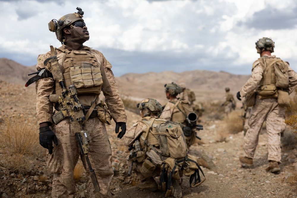 A group of military personnel in desert camouflage uniforms are conducting operations in an arid, mountainous terrain. The foreground soldier is looking upward while holding a rifle, with additional personnel visible in various positions behind him. The setting appears to be a desert training or operational environment with rocky hills in the background.