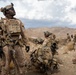 A group of military personnel in desert camouflage uniforms are conducting operations in an arid, mountainous terrain. The foreground soldier is looking upward while holding a rifle, with additional personnel visible in various positions behind him. The setting appears to be a desert training or operational environment with rocky hills in the background.