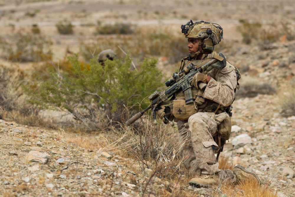 A heavily equipped military service member in desert camouflage (MARPAT pattern) is kneeling in a rocky, arid terrain while holding an assault rifle with an optic scope. A second service member is partially visible in the background behind desert scrub brush. The setting appears to be a desert training environment or operational area.