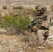 A heavily equipped military service member in desert camouflage (MARPAT pattern) is kneeling in a rocky, arid terrain while holding an assault rifle with an optic scope. A second service member is partially visible in the background behind desert scrub brush. The setting appears to be a desert training environment or operational area.