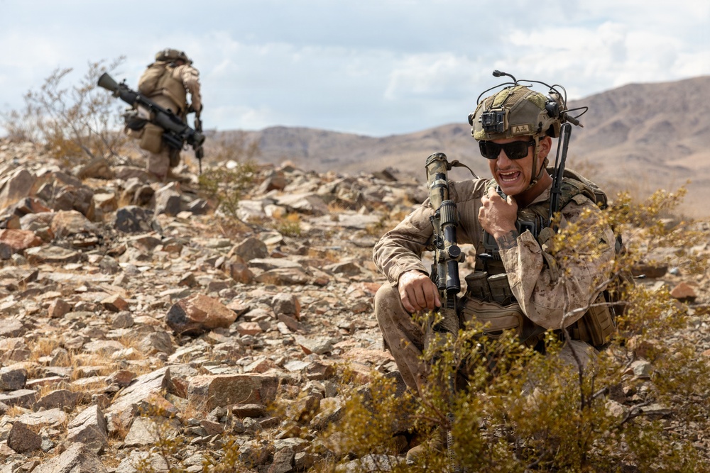 Two military personnel in desert camouflage uniforms are operating in a rocky, arid terrain with mountains visible in the background. The foreground soldier is crouching and appears to be communicating via radio while holding a rifle with an optic scope. The background soldier is moving forward while carrying a weapon, suggesting a patrol or training exercise.