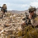 Two military personnel in desert camouflage uniforms are operating in a rocky, arid terrain with mountains visible in the background. The foreground soldier is crouching and appears to be communicating via radio while holding a rifle with an optic scope. The background soldier is moving forward while carrying a weapon, suggesting a patrol or training exercise.