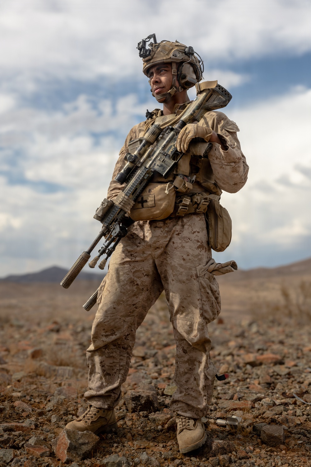 A fully equipped military service member stands in an arid, rocky desert terrain while holding a rifle across their chest. The soldier is wearing desert camouflage (MARPAT pattern consistent with USMC), tactical helmet with night-vision mount, and full combat gear. The image appears to be a professional or official military photograph given its high production quality.