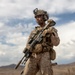 A fully equipped military service member stands in an arid, rocky desert terrain while holding a rifle across their chest. The soldier is wearing desert camouflage (MARPAT pattern consistent with USMC), tactical helmet with night-vision mount, and full combat gear. The image appears to be a professional or official military photograph given its high production quality.