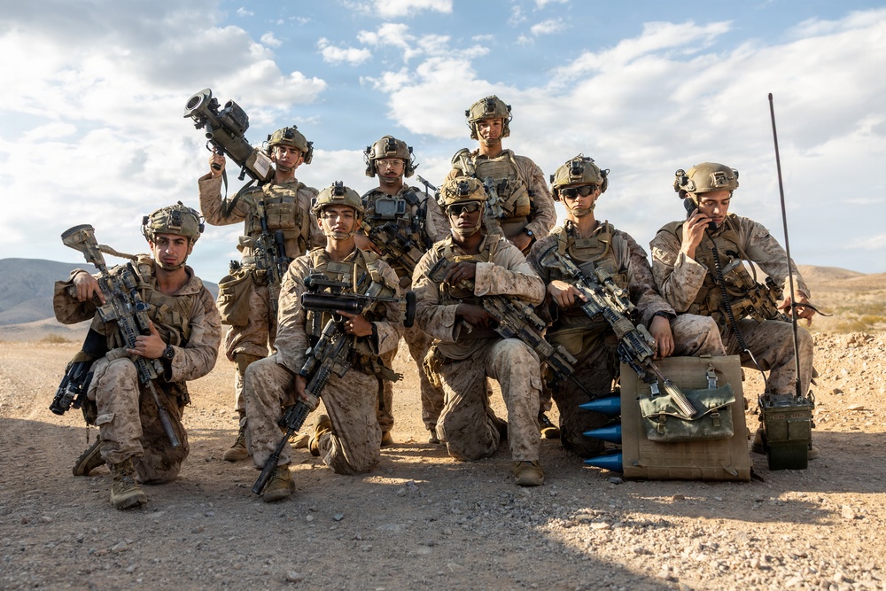 A group of approximately nine military personnel in desert camouflage uniforms pose together in an arid, desert environment. They are fully equipped with tactical gear, weapons, helmets, and communication equipment. The image appears to be a unit group photo taken during a field exercise or deployment.