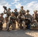 A group of approximately nine military personnel in desert camouflage uniforms pose together in an arid, desert environment. They are fully equipped with tactical gear, weapons, helmets, and communication equipment. The image appears to be a unit group photo taken during a field exercise or deployment.