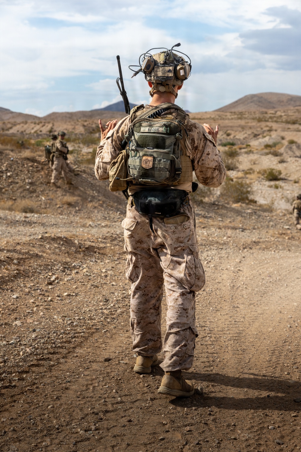 A U.S. Marine, identified by the name tape 'NORRIS' on their pack, is seen from behind walking along a dirt trail in an arid desert environment. The Marine is in full combat gear including MARPAT desert camouflage, a tactical helmet with communications equipment, a radio antenna, and a tactical backpack. At least one additional Marine is visible in the background, suggesting a patrol or field training exercise.