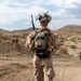 A U.S. Marine, identified by the name tape 'NORRIS' on their pack, is seen from behind walking along a dirt trail in an arid desert environment. The Marine is in full combat gear including MARPAT desert camouflage, a tactical helmet with communications equipment, a radio antenna, and a tactical backpack. At least one additional Marine is visible in the background, suggesting a patrol or field training exercise.
