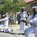A group of U.S. Navy personnel in dress white uniforms are performing a ceremonial rifle drill in an outdoor plaza area. The drill team members are executing synchronized movements with rifles equipped with bayonets, with some kneeling and others standing. The performance appears to be taking place in front of a historic stone building, likely during a public ceremony or demonstration.