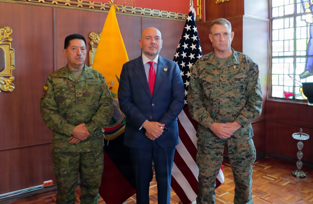 Three individuals pose formally in what appears to be an official meeting room or government building. Two are wearing military uniforms — one in Ecuadorian Army camouflage and one in U.S. Marine Corps MARPAT camouflage — while the central figure wears a civilian business suit with a red tie. The flags of Ecuador and the United States are visible in the background, suggesting a bilateral military or diplomatic engagement.