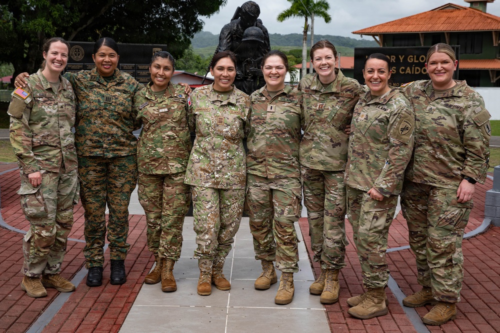 Eight female military personnel in various camouflage uniforms pose together for a group photo at what appears to be a military installation in a Latin American country. The group includes soldiers from multiple nations, identifiable by different uniform patterns including US Army OCP, and what appears to be a Marine Corps MARPAT uniform. They are standing in front of a bronze military memorial statue on a brick plaza.