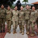 Eight female military personnel in various camouflage uniforms pose together for a group photo at what appears to be a military installation in a Latin American country. The group includes soldiers from multiple nations, identifiable by different uniform patterns including US Army OCP, and what appears to be a Marine Corps MARPAT uniform. They are standing in front of a bronze military memorial statue on a brick plaza.