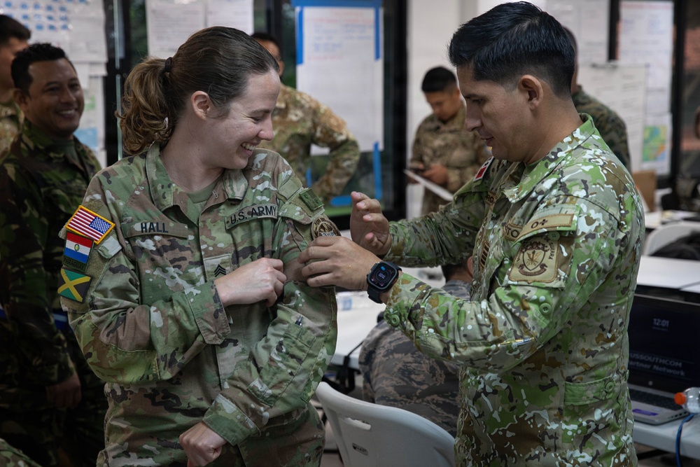 A U.S. Army soldier named Hall is receiving what appears to be a military coin or badge from a foreign military officer in a multinational military setting. The interaction is friendly and ceremonial, with both individuals smiling. The background shows additional military personnel from various nations in what appears to be a command or operations center.