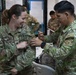 A U.S. Army soldier named Hall is receiving what appears to be a military coin or badge from a foreign military officer in a multinational military setting. The interaction is friendly and ceremonial, with both individuals smiling. The background shows additional military personnel from various nations in what appears to be a command or operations center.