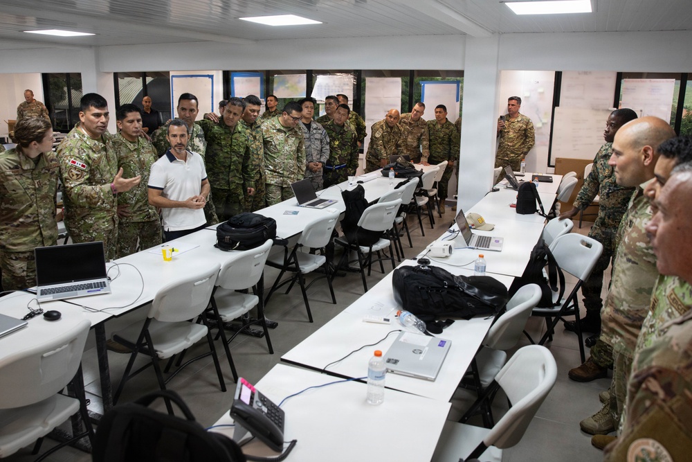 A group of military personnel in various camouflage uniforms are gathered in a briefing or training room, appearing to receive instruction or participate in a discussion. The room contains multiple white folding tables arranged in rows with laptops, bags, and other equipment on them. One individual in civilian clothing appears to be presenting or speaking to the assembled group.
