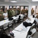 A group of military personnel in various camouflage uniforms are gathered in a briefing or training room, appearing to receive instruction or participate in a discussion. The room contains multiple white folding tables arranged in rows with laptops, bags, and other equipment on them. One individual in civilian clothing appears to be presenting or speaking to the assembled group.