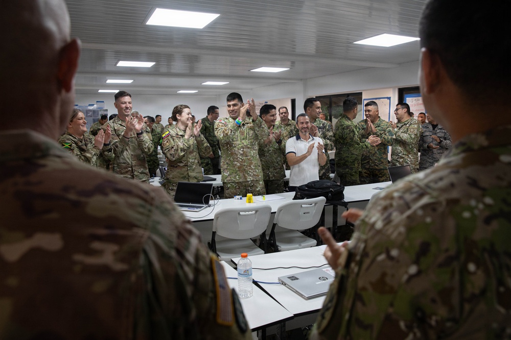 A group of military personnel from multiple nations are gathered in a classroom or briefing room, applauding in what appears to be a multinational training or conference event. The participants are wearing various camouflage uniforms, with at least one individual in civilian attire. The atmosphere is positive and celebratory, suggesting a conclusion of a session or recognition moment.