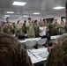 A group of military personnel from multiple nations are gathered in a classroom or briefing room, applauding in what appears to be a multinational training or conference event. The participants are wearing various camouflage uniforms, with at least one individual in civilian attire. The atmosphere is positive and celebratory, suggesting a conclusion of a session or recognition moment.
