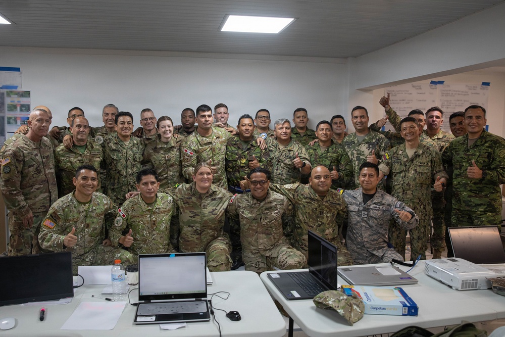 A large group of military personnel from multiple nations pose together for a group photo in what appears to be a briefing or operations room. The group includes both male and female soldiers wearing various camouflage uniforms with different national patches and insignia. The setting suggests a multinational military exercise, training event, or joint operations meeting.