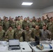 A large group of military personnel from multiple nations pose together for a group photo in what appears to be a briefing or operations room. The group includes both male and female soldiers wearing various camouflage uniforms with different national patches and insignia. The setting suggests a multinational military exercise, training event, or joint operations meeting.