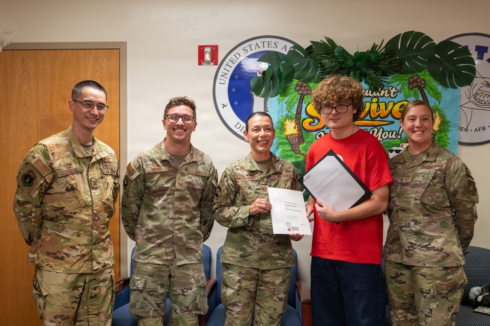 Four U.S. Air Force personnel in OCP (Operational Camouflage Pattern) uniforms pose with a civilian teenager in a red shirt inside a military facility. A certificate presentation is taking place, with what appears to be a Red Cross Certificate of Appreciation being held jointly by a female service member and the young civilian. The setting appears to be at an Air Force Base, as indicated by the United States Air Force seal and 'AFB' text visible on the wall.