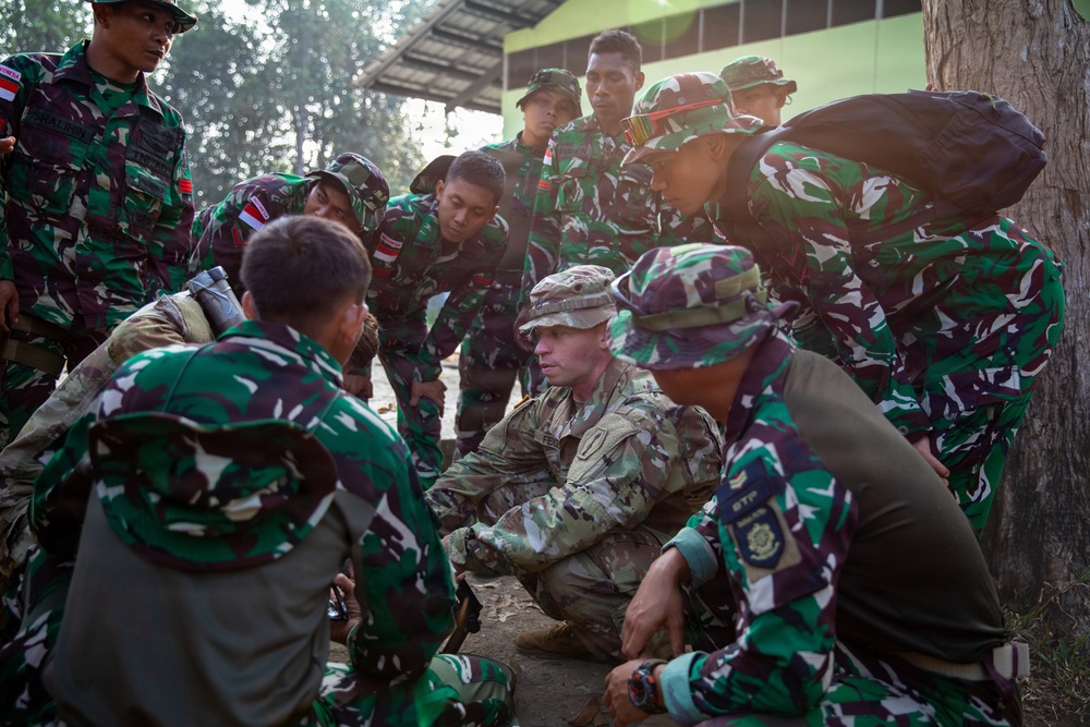 A group of soldiers in green camouflage uniforms, appearing to be Indonesian military (TNI - Tentara Nasional Indonesia), are gathered around one soldier in a different camouflage pattern (likely US Army MultiCam/OCP uniform) who appears to be conducting a training or briefing session. The group is crouched or standing in a tight circle, focused on something at ground level, suggesting a field training exercise or tactical instruction. The setting appears to be an outdoor military training area.