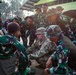 A group of soldiers in green camouflage uniforms, appearing to be Indonesian military (TNI - Tentara Nasional Indonesia), are gathered around one soldier in a different camouflage pattern (likely US Army MultiCam/OCP uniform) who appears to be conducting a training or briefing session. The group is crouched or standing in a tight circle, focused on something at ground level, suggesting a field training exercise or tactical instruction. The setting appears to be an outdoor military training area.