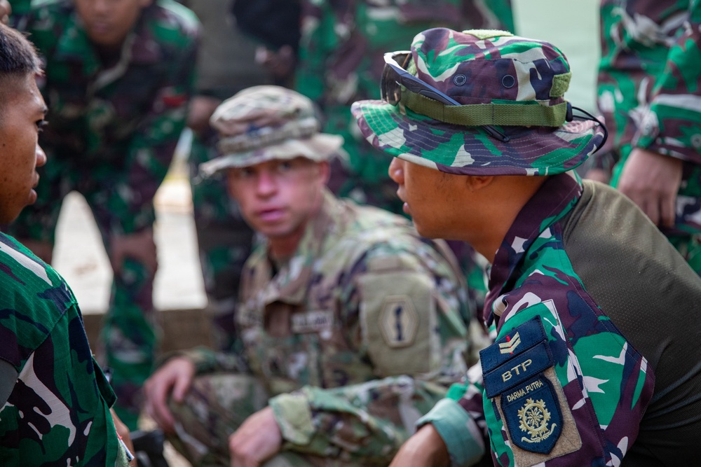 The image shows a joint military training or coordination session involving at least one U.S. Army soldier and several Indonesian Armed Forces (TNI) personnel. The U.S. soldier wears MultiCam (OCP) uniform with a '1st Infantry Division' patch, while the Indonesian soldiers wear their distinctive green camouflage (loreng) uniforms. The foreground Indonesian soldier displays a 'BTP / DARMA PUTRA' unit patch, indicating a specific TNI battalion affiliation.