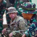 The image shows a joint military training or coordination session involving at least one U.S. Army soldier and several Indonesian Armed Forces (TNI) personnel. The U.S. soldier wears MultiCam (OCP) uniform with a '1st Infantry Division' patch, while the Indonesian soldiers wear their distinctive green camouflage (loreng) uniforms. The foreground Indonesian soldier displays a 'BTP / DARMA PUTRA' unit patch, indicating a specific TNI battalion affiliation.