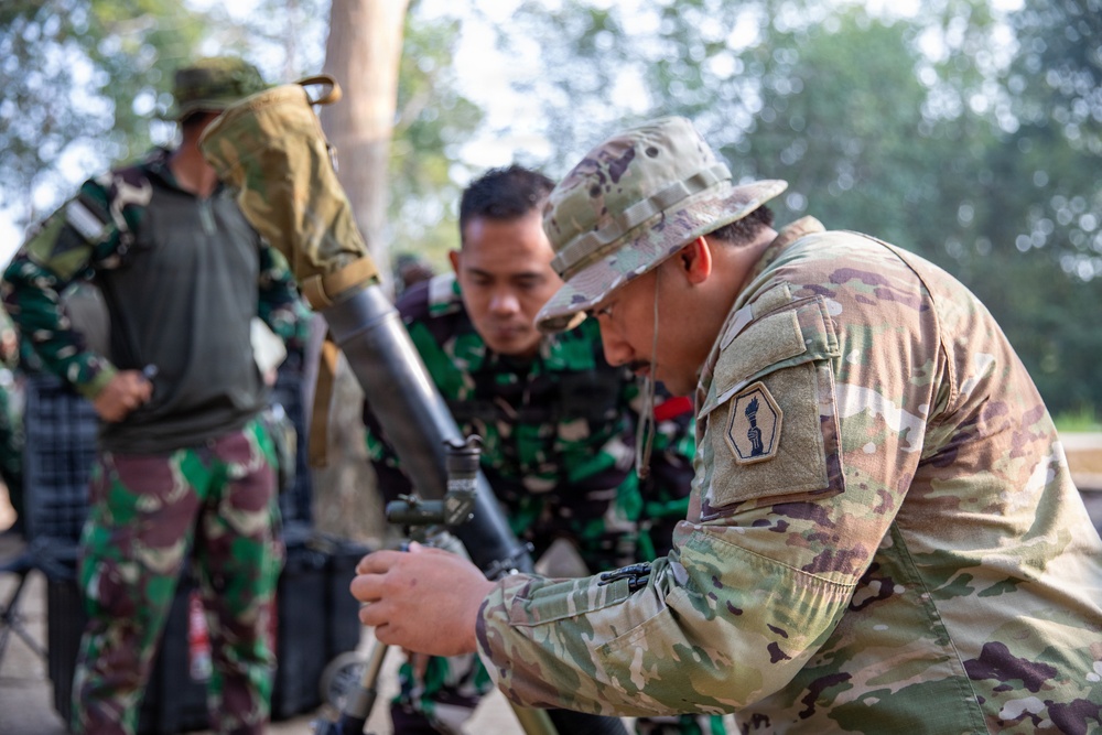 The image shows military personnel from at least two different armed forces working together around what appears to be a mortar system. A soldier in MultiCam/OCP uniform with a US Army patch is closely examining or adjusting the mortar alongside soldiers wearing Indonesian TNI (Tentara Nasional Indonesia) camouflage uniforms. The setting appears to be an outdoor training environment, suggesting a joint military exercise or training event.