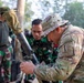 The image shows military personnel from at least two different armed forces working together around what appears to be a mortar system. A soldier in MultiCam/OCP uniform with a US Army patch is closely examining or adjusting the mortar alongside soldiers wearing Indonesian TNI (Tentara Nasional Indonesia) camouflage uniforms. The setting appears to be an outdoor training environment, suggesting a joint military exercise or training event.
