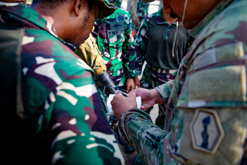 Military personnel from at least two different armed forces are gathered in close proximity, appearing to review notes or documentation. One soldier wears Indonesian Armed Forces (TNI) camouflage and another wears US Army MultiCam (OCP) uniform with a visible unit patch. The interaction suggests a joint training exercise or coordination meeting between allied military forces.